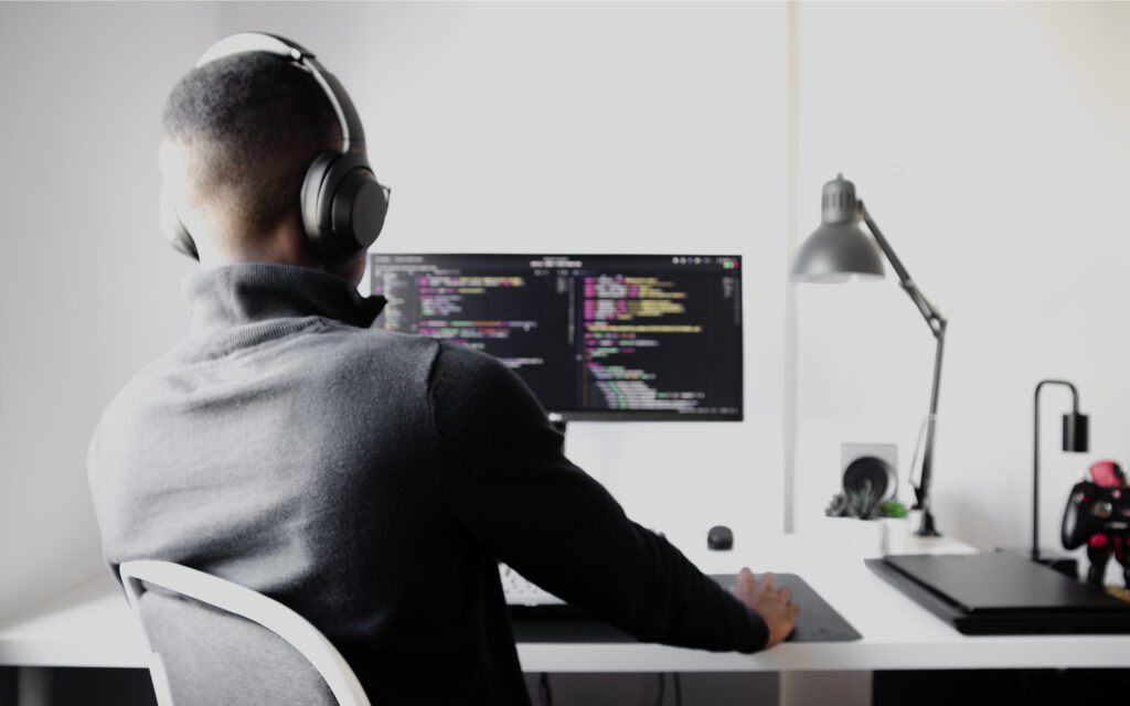 Junger Mann mit Kopfhörern sitzt am Schreibtisch vor einem Bildschirm voller Code